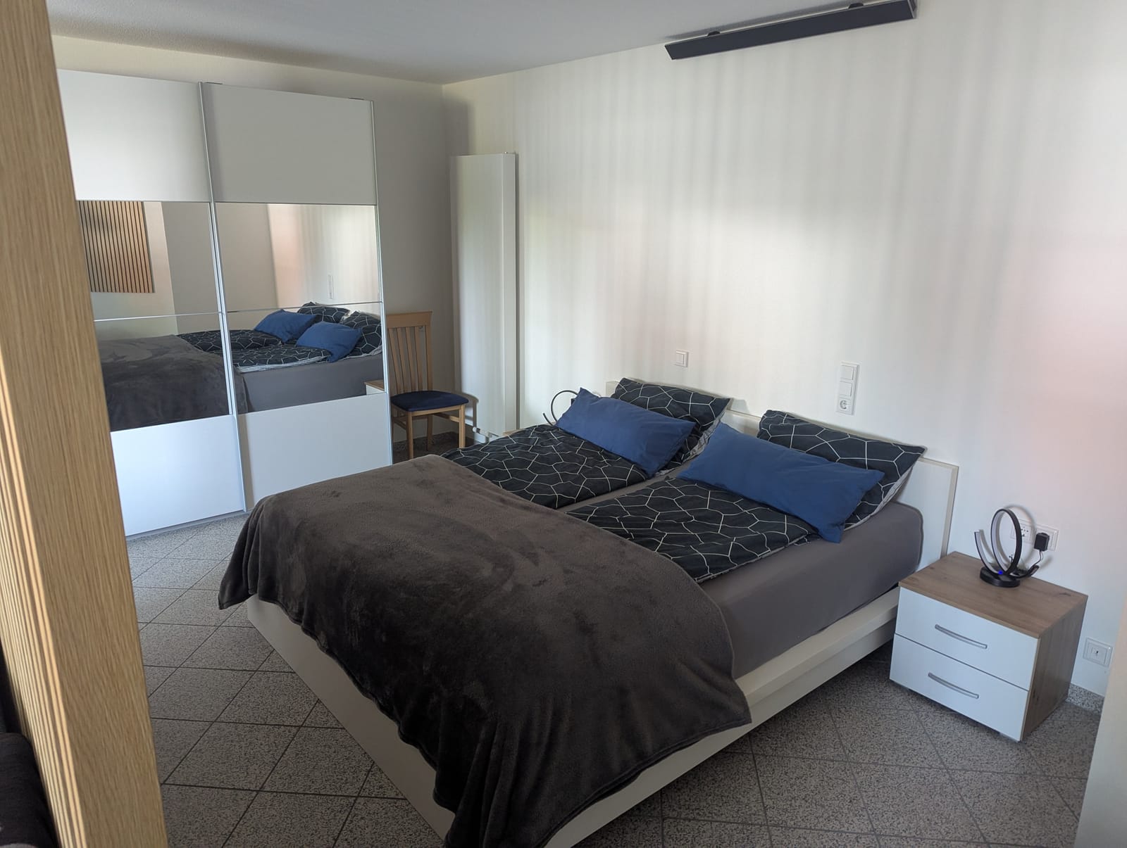 Schlafbereich mit Doppelbett, Nachttisch und großem Kleiderschrank mit Spiegeltüren, abgetrennt durch eine Holzlamellenwand