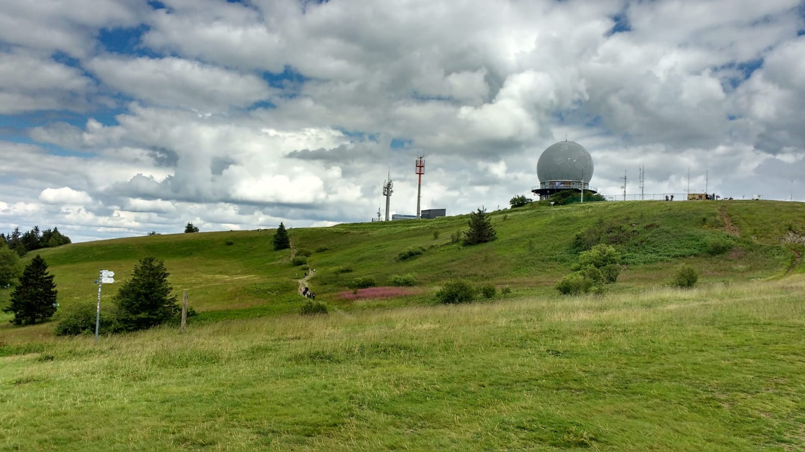 Wanderer auf einem Wiesenweg zur Wasserkuppe, oben die Radarkuppel unter Sommerwolken