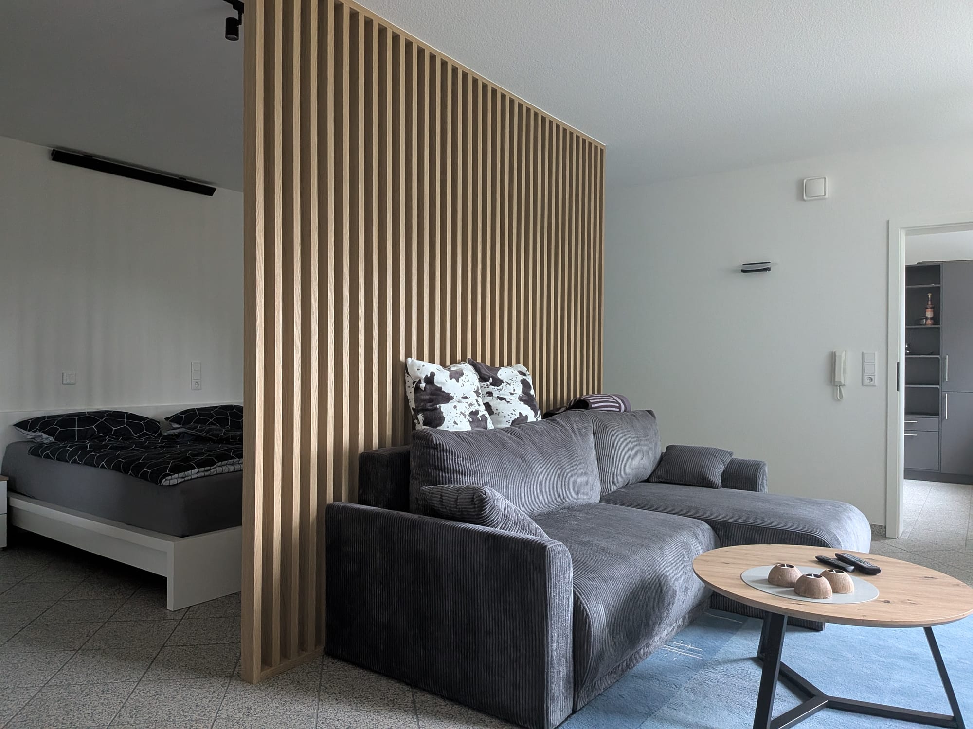 Der offene Wohn-/Schlafraum: links das Doppelbett, rechts die Couchgarnitur mit Couchtisch, dazwischen eine raumhohe Holzlamellenwand als Raumteiler. Decke und Fliesenboden laufen durch